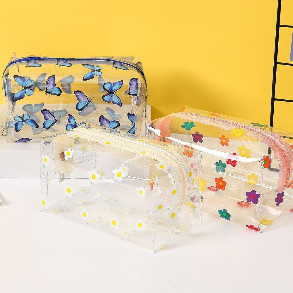 Three clear plastic zippered cosmetic bags with different prints: blue butterflies, white daisies with yellow centers, and multicolored flowers, displayed on a white surface against a yellow background.