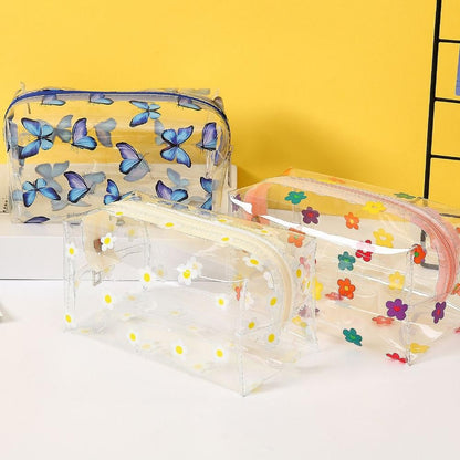 Three clear plastic zippered cosmetic bags with different prints: blue butterflies, white daisies with yellow centers, and multicolored flowers, displayed on a white surface against a yellow background.