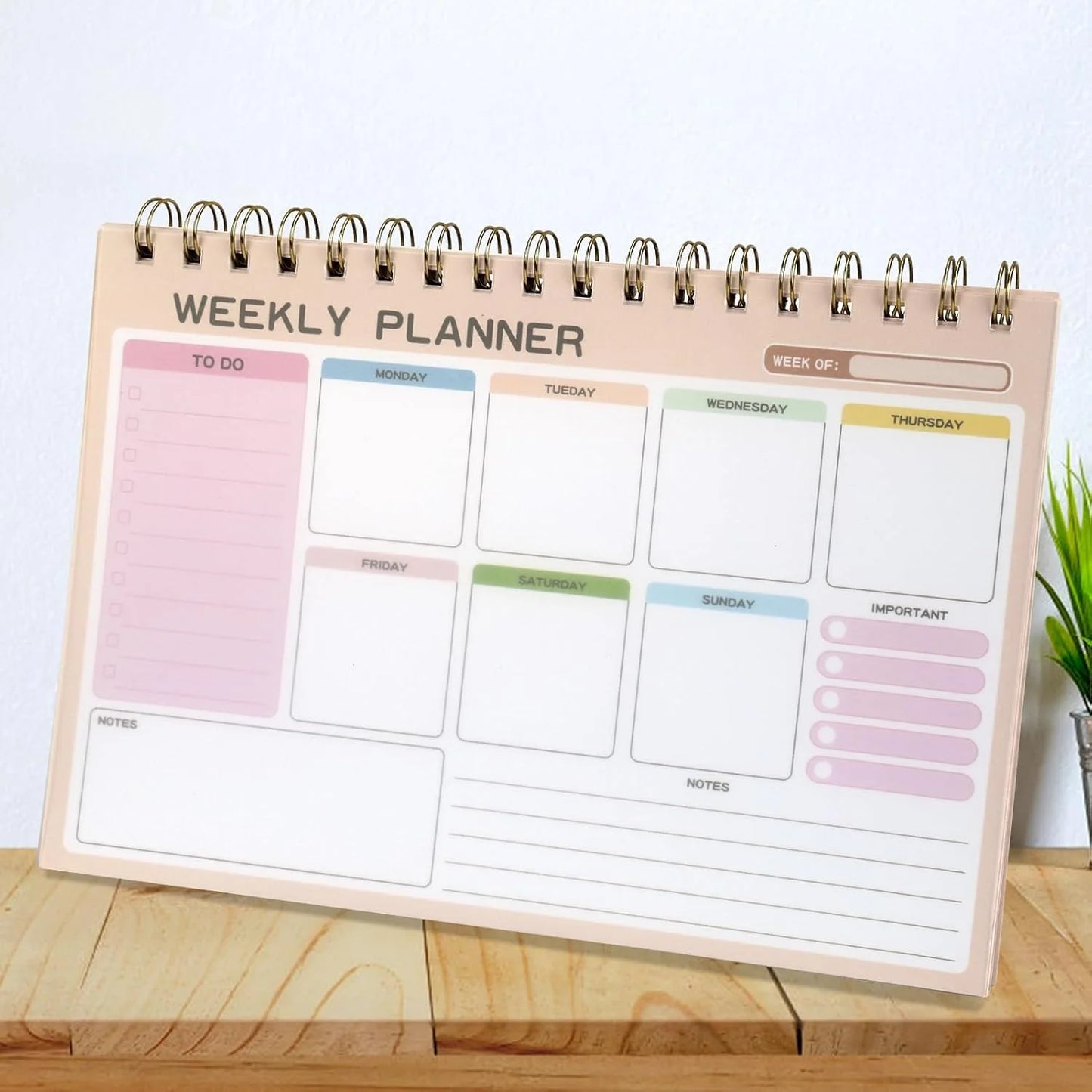 A spiral-bound weekly planner notepad displayed on a wooden surface against a white wall. The planner has a light peach-beige cover with sections for to-do, daily tasks from Monday to Sunday, an important notes column, and a week overview at the top.