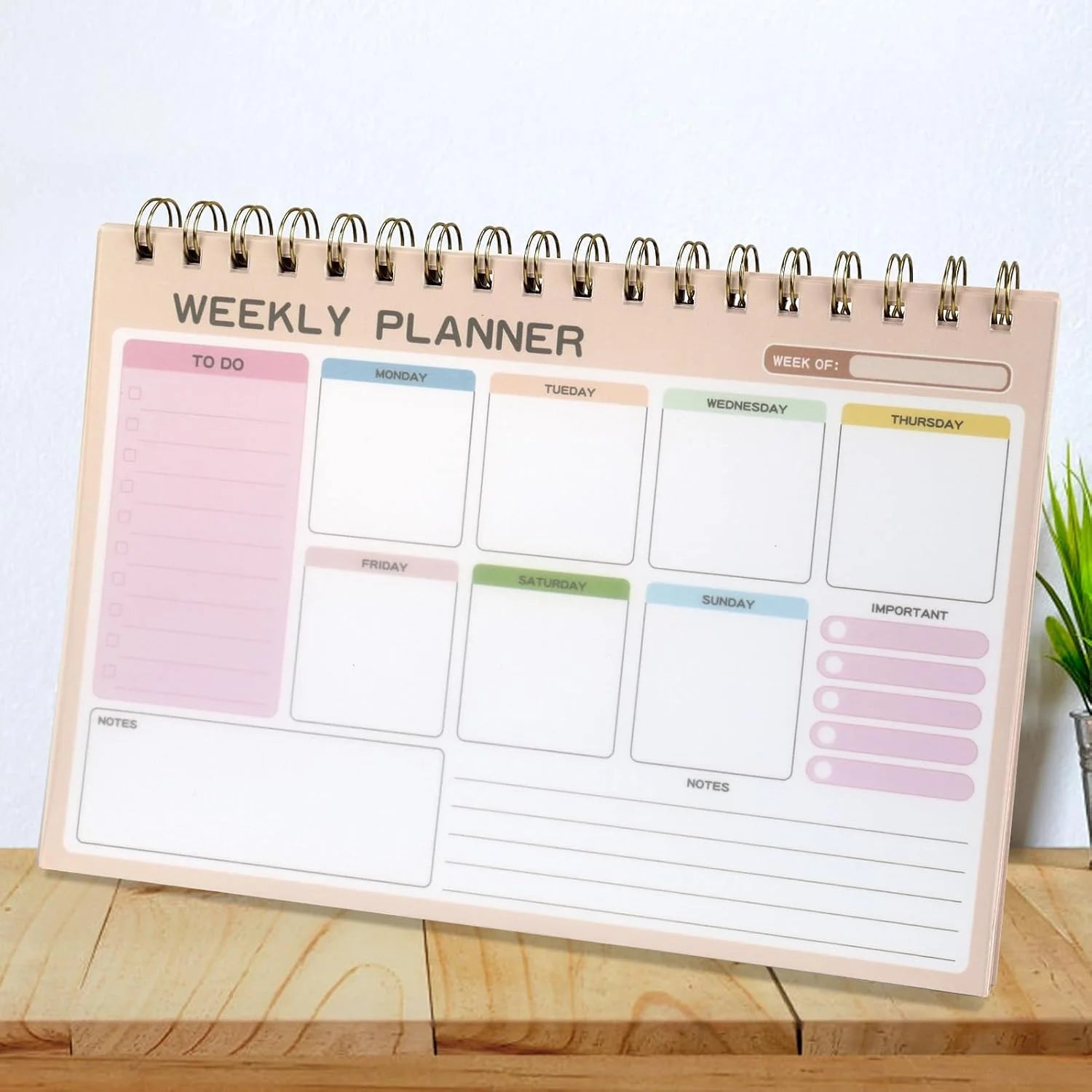 A spiral-bound weekly planner notepad displayed on a wooden surface against a white wall. The planner has a light peach-beige cover with sections for to-do, daily tasks from Monday to Sunday, an important notes column, and a week overview at the top.