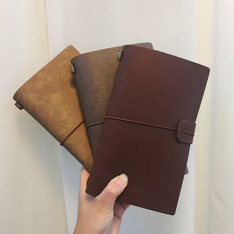 Three leather-bound travel journals in brown, black, and red colors, held in a hand with a visible elastic closure on the brown journal.